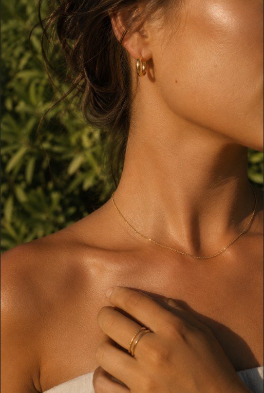 a close crop on a sun tanned neck and shoulder wearing a fine gold chain and a small gold hoop earring, set outdoors against soft green foliage, with warm late afternoon light skimming the skin, quiet and intimate mood, and details including a couple of thin gold rings on the hand resting near the collarbone.