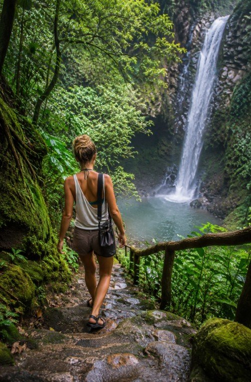 a woman in shorts and a light top with a swimsuit visible underneath, walking down mossy stone steps toward a jungle waterfall, set deep in dense green forest, with cool filtered light through the canopy and mist rising from below, adventurous and grounded mood, and details including a small dry bag on her shoulder and grippy sandals on wet stone.
