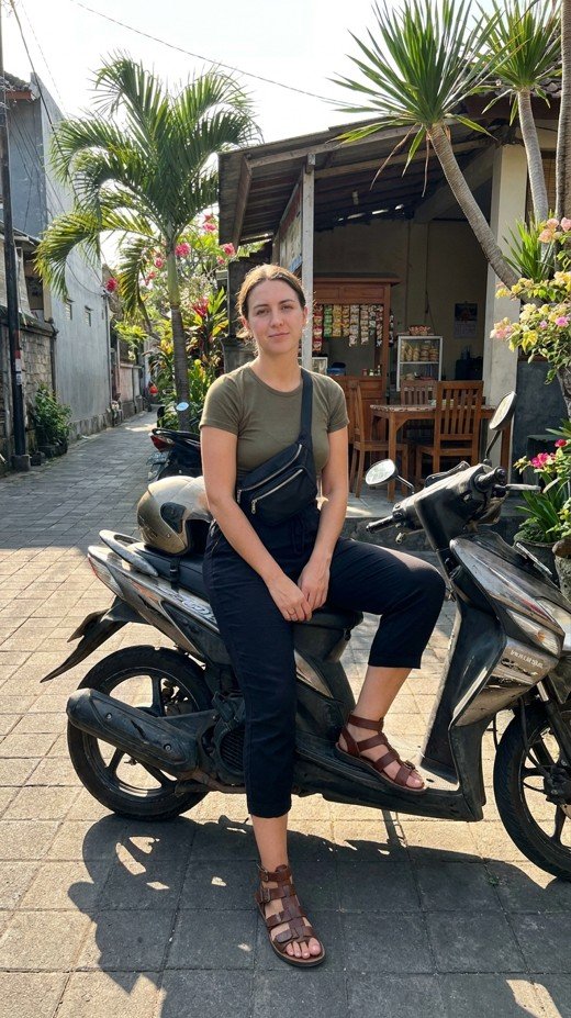a woman in a dark cropped trouser and a fitted top sitting on a parked scooter at the roadside, set on a narrow Bali street with a warung and greenery behind, with bright hard afternoon sun and crisp shadow, practical and everyday mood, and details including a zipped crossbody worn across the chest, secure strapped sandals, and helmet resting on the seat.